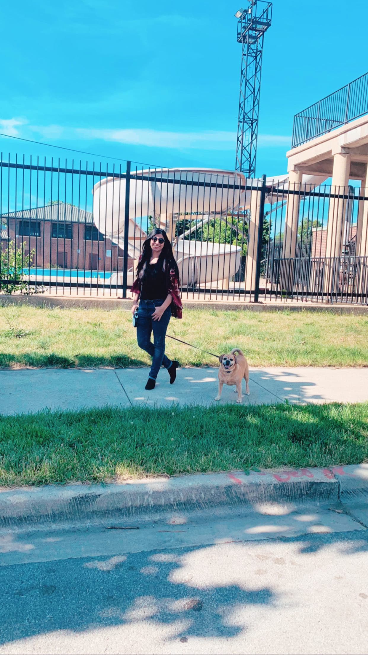 Woman walks a small tan dog on a sidewalk past a large white water slide.